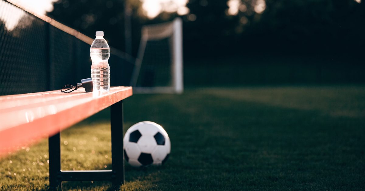 A soccer ball on the grass beside a sideline bench with a water bottle and a coach's whistle, on an empty youth soccer field at dusk.