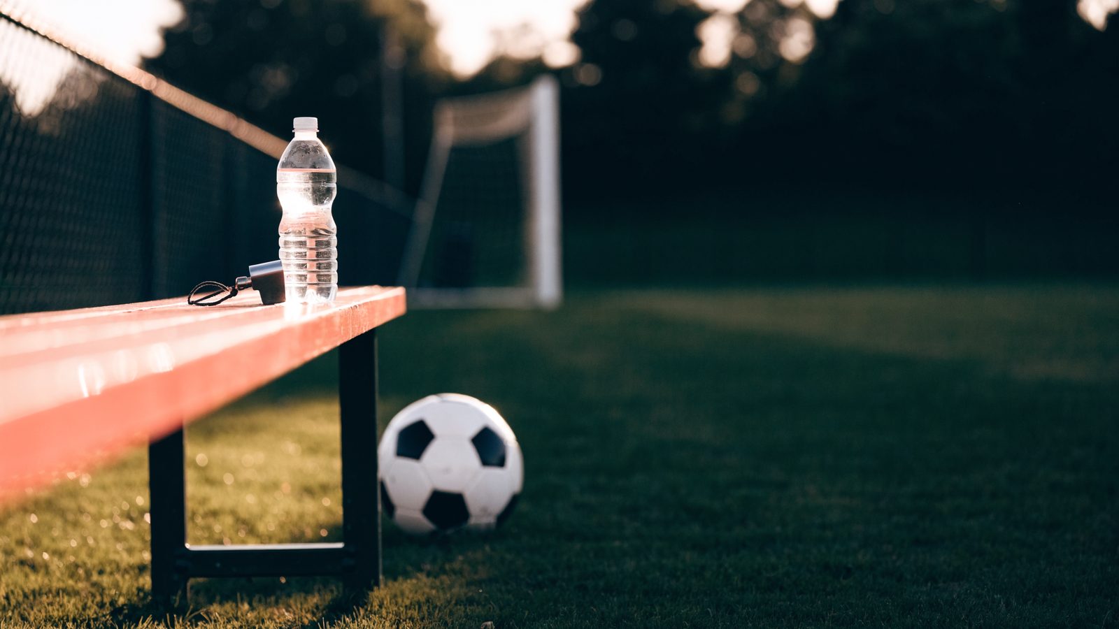A soccer ball on the grass beside a sideline bench with a water bottle and a coach's whistle, on an empty youth soccer field at dusk.